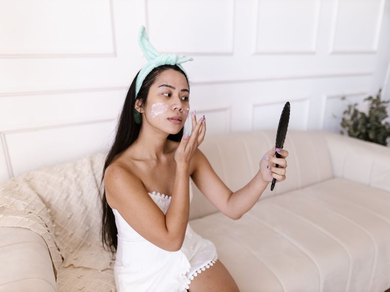 Phụ nữ châu Á thoa kem dưỡng chứa hyaluronic acid và kiểm tra da qua gương
